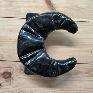 Pode incluir: Uma ferramenta de modelagem de cabelo em forma de crescente preto com uma superfície texturizada. A ferramenta é montada em uma base preta.