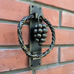 May include: A black metal door knocker with a grape cluster design. The knocker is attached to a brick wall.