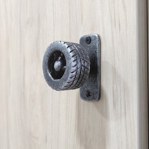 Könnte beinhalten: Ein silberner Metall-Schranktürknopf mit einem Reifenprofil-Design. Der Knopf ist auf einer rechteckigen Platte montiert.