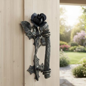 Könnte beinhalten: Ein schwarzer Metalltürgriff mit Rosenmotiv. Der Griff zeigt eine detaillierte Rosenblüte, Blätter und einen strukturierten, astähnlichen Griff. Der Griff ist an einer hellen Holztür montiert, mit einer verschwommenen Szene im Freien im Hintergrund.