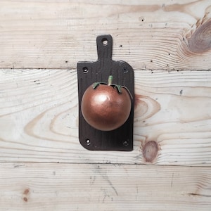 May include: A brown metal cutting board with a copper-colored tomato attached to the front. The cutting board has a handle and a textured surface.