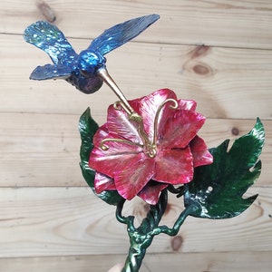 May include: A metal sculpture of a hummingbird hovering over a red flower with green leaves. The hummingbird is blue and green with a long beak. The flower is a hibiscus with five petals.