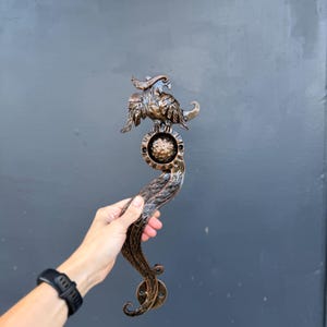 May include: A bronze-colored metal door handle featuring a sculpted bird figure at the top, a central circular plate with a textured sphere, and a long, curved, flowing tail design that ends in a circular mounting base with two screw holes.