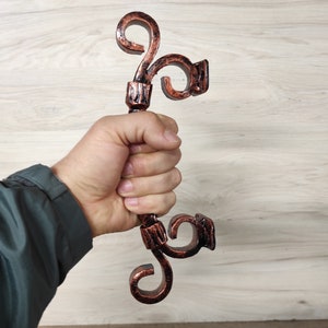 Op de afbeelding: Een hand houdt een decoratieve, sierlijke, koperkleurige metalen handgreep vast met een zwierig ontwerp. De handgreep is waarschijnlijk voor een deur of kast.