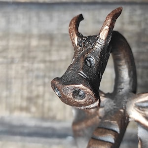 May include: Close-up of a dragon sculpture with a copper finish. The dragon's head is facing forward with its mouth closed. The dragon's eyes are black and the horns are pointed.