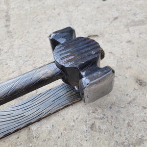Puede incluir: Primer plano de un martillo de metal negro con mango y cabeza texturizados. La cabeza del martillo tiene una superficie de golpe plana y una parte superior redondeada con ranuras horizontales. El mango y la base tambi&eacute;n est&aacute;n texturizados.