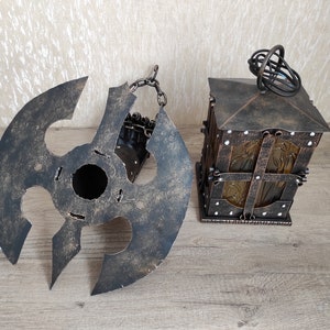 May include: A rustic metal lantern with a decorative glass panel and a chain hanging from the top. The lantern is in a dark brown colour with a distressed finish. The lantern is sitting on a wooden surface.