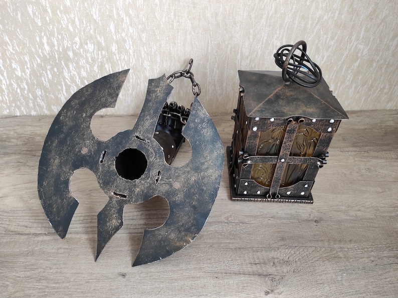 May include: A rustic metal lantern with a decorative glass panel and a chain hanging from the top. The lantern is in a dark brown colour with a distressed finish. The lantern is sitting on a wooden surface.