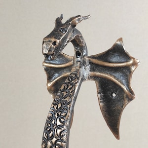 May include: A black and gold dragon door knocker with intricate details. The dragon's wings are spread out and the head is facing forward. The knocker is made of metal and has a rustic finish.