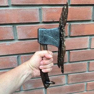 Peut inclure: Une main tient une hache noire et brune avec un motif métallique décoratif. La hache a un anneau à l'extrémité du manche.