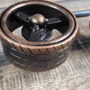 May include: A close-up of a decorative knob with a black tire tread design and a copper-colored rim. The knob features a three-spoke wheel design with a gold-colored center hub.