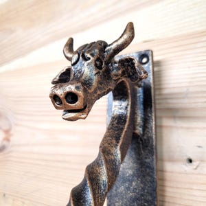 May include: A bronze-colored metal cow head door handle. The cow head is detailed with horns, eyes, and a smiling mouth. The handle has a twisted design and is mounted on a rectangular plate.