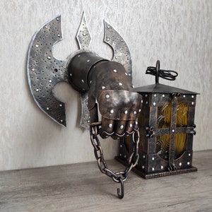 May include: A metal wall sconce featuring a hand-shaped design holding a small lantern. The hand is clad in armour and is holding a chain. The lantern is made of metal and has a glass window. The sconce is painted in a dark brown colour.