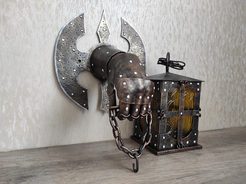 May include: A metal wall sconce featuring a hand-shaped design holding a small lantern. The hand is clad in armour and is holding a chain. The lantern is made of metal and has a glass window. The sconce is painted in a dark brown colour.