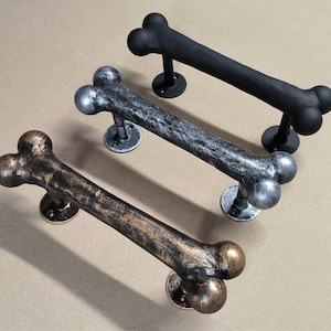 Puede incluir: Tres tiradores de puerta con forma de hueso en negro, plateado y bronce. Cada tirador tiene una base redonda para el montaje y está hecho de metal. Los tiradores están diseñados para parecerse a un hueso de perro.