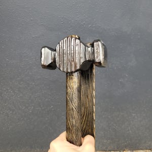 Puede incluir: Un martillo artesanal con una cabeza met&aacute;lica oscura y un mango de madera. La cabeza del martillo presenta una superficie texturizada y el mango est&aacute; tallado con un patr&oacute;n de vetas de madera. El martillo se sostiene en una mano.