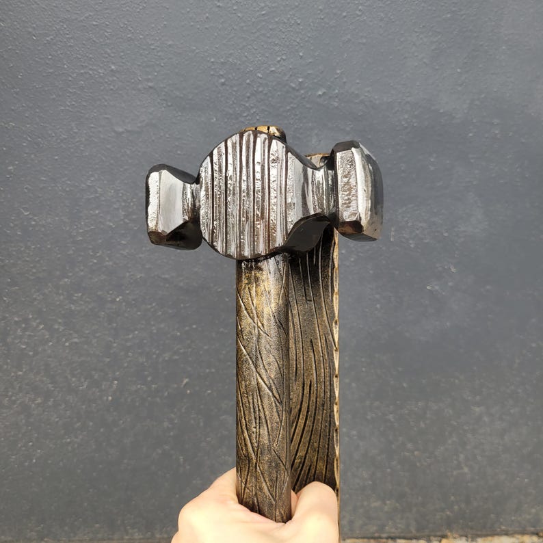 Puede incluir: Un martillo artesanal con una cabeza met&aacute;lica oscura y un mango de madera. La cabeza del martillo presenta una superficie texturizada y el mango est&aacute; tallado con un patr&oacute;n de vetas de madera. El martillo se sostiene en una mano.