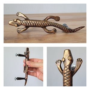 Puede incluir: Tirador de armario con forma de lagarto de color bronce, con cuerpo texturizado y patas y cola detalladas. El tirador del armario está montado con dos tornillos. El lagarto está sobre una superficie de madera.