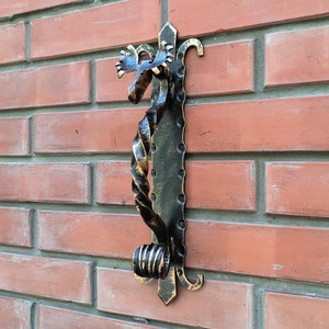 Puede incluir: Un llamador de puerta de metal negro con un diseño retorcido y una cabeza de alce en la parte superior. El llamador está montado en una pared de ladrillo.