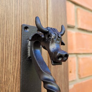 May include: Black cast iron door knocker featuring a cow head design. The knocker is mounted on a rectangular plate and attached to a wooden door. The handle has a twisted design.