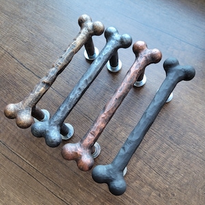 Può includere: Quattro maniglie per porte a forma di osso in bronzo, grigio, rame e nero. Ogni maniglia è montata con una base e una vite color argento. Le maniglie hanno un aspetto strutturato e martellato, aggiungendo un tocco rustico e unico a qualsiasi porta o armadio.