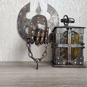 May include: A decorative metal wall hanging featuring a hand in a chainmail glove, attached to a chain and a wall-mounted axe. A small, ornate metal lantern with a glass panel is also visible.