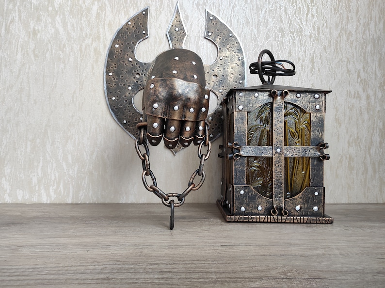 May include: A decorative metal wall hanging featuring a hand in a chainmail glove, attached to a chain and a wall-mounted axe. A small, ornate metal lantern with a glass panel is also visible.