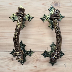 May include: Two ornate door handles with a rustic, twig-like design. The handles are made of metal with a gold finish and have a green patina. The handles are attached to a wooden surface.