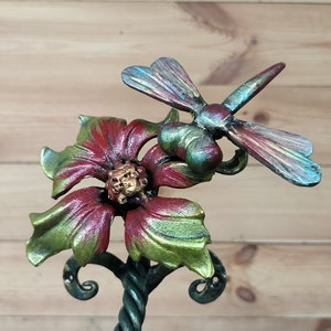 May include: A metal sculpture of a flower with a dragonfly perched on top. The flower is red and green with a gold center. The dragonfly is iridescent blue and green.