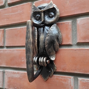 May include: A black metal owl door knocker with a gold finish. The owl is perched on a branch with its wings spread out. The knocker is mounted on a brick wall.