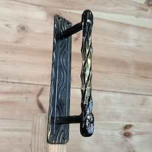 May include: Black metal door handle with a decorative, twisted design. The handle has a rectangular backplate with a wavy pattern.