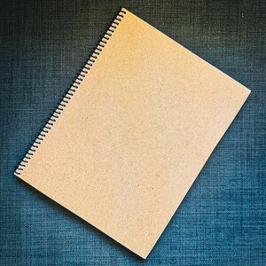 K&ouml;nnte beinhalten: Ein Spiralnotizbuch mit einem Einband aus Kraftpapier. Das Notizbuch ist ge&ouml;ffnet und zeigt leere Seiten. Die Bindung ist schwarz, und das Notizbuch liegt auf einem strukturierten, blauen Hintergrund. Das Design ist schlicht und funktional.