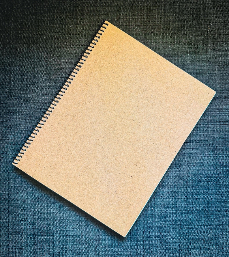 K&ouml;nnte beinhalten: Ein Spiralnotizbuch mit einem Einband aus Kraftpapier. Das Notizbuch ist ge&ouml;ffnet und zeigt leere Seiten. Die Bindung ist schwarz, und das Notizbuch liegt auf einem strukturierten, blauen Hintergrund. Das Design ist schlicht und funktional.