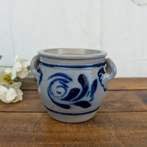 May include: A gray stoneware crock featuring two side handles and a dark blue hand-painted floral swirl pattern. The cylindrical vessel has a thick rim and a smooth glazed finish, showcasing a classic salt-glaze style with deep blue cobalt accents.