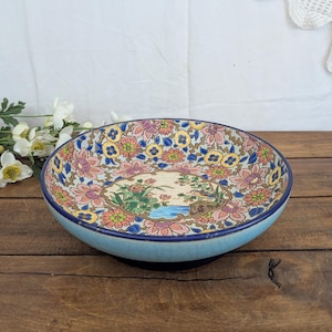 Op de afbeelding: Een ronde keramische kom met een lichtblauwe buitenkant en een donkerblauwe rand. De binnenzijde toont een centraal medaillon met een vogel bij een vijver, omringd door een dicht patroon van roze, gele en groene bloemmotieven op een crèmekleurige achtergrond.