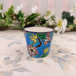 May include: A small blue ceramic cup with a floral pattern. The cup has a white interior and a blue rim. The floral pattern features pink and yellow flowers, green leaves, and brown branches.