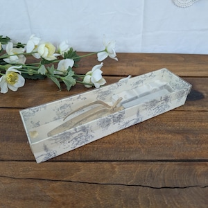 Puede incluir: Una caja de regalo rectangular con tapa transparente, que contiene un par de utensilios de servir plateados. La caja tiene un fondo crema con un patrón azul y gris. La caja está sobre una superficie de madera, con flores blancas y vegetación en el fondo.