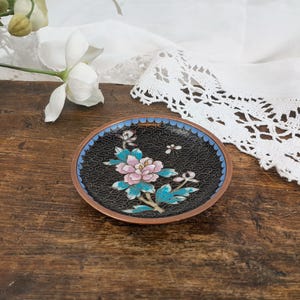 May include: An oval, decorative dish with a black background and a floral design. The dish has a copper-colored rim and a blue border. The floral design features pink and blue flowers with green leaves. The dish is on a wooden surface.