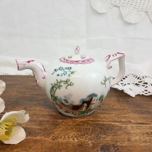 Peut inclure: Une théière en porcelaine blanche au corps arrondi, décorée d'un paysage et d'éléments floraux. Le couvercle et le bec sont ornés de détails roses et violets. La théière est posée sur une surface en bois.