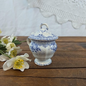 May include: A blue and white ceramic sugar bowl with a lid, decorated with a floral pattern. The bowl has a pedestal base and a small handle on the lid. The background includes white flowers and a wooden surface.