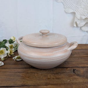 Peut inclure: Un plat de service en céramique marron clair avec couvercle et deux poignées. Le couvercle a un petit bouton et un motif de cercles concentriques. Le plat est sur une surface en bois avec des fleurs blanches.