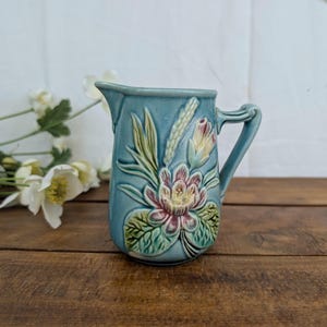 Peut inclure: Un pichet en céramique bleu clair orné d'un motif floral en relief avec des feuilles vertes, des pétales de fleurs roses et blancs, et un épi de blé. Le pichet présente une anse incurvée et un bec verseur avec une finition brillante.