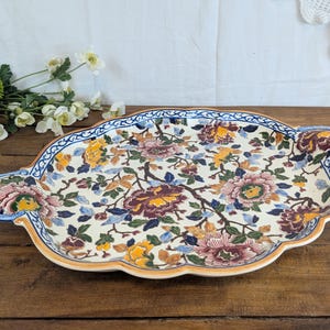 May include: An oval ceramic serving platter with a scalloped edge, featuring a cream-colored background decorated with a detailed floral pattern in shades of deep red, yellow, green, and blue, finished with a blue and white decorative border.