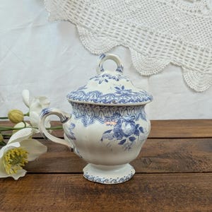 Peut inclure: Un sucrier en céramique blanc et bleu avec un couvercle. Le sucrier présente un motif floral et une poignée sur le côté. Le couvercle a une petite poignée sur le dessus. Le sucrier est sur une surface en bois.