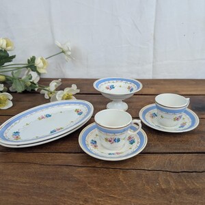 Puede incluir: Un juego de dos tazas de té de porcelana blanca con sus platos, un pequeño cuenco con pedestal y dos platos ovalados. Cada pieza presenta un borde azul claro, detalles dorados y un delicado estampado floral con flores rosas y azules.
