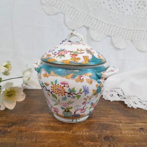 Puede incluir: Un cuenco de cerámica vintage con tapa y diseño floral. El cuenco presenta una banda turquesa, detalles dorados y un fondo blanco con flores y follaje coloridos. La tapa tiene un pequeño asa y un patrón floral a juego.