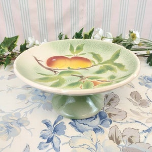 Peut inclure: Un support à gâteau en céramique vert et blanc avec un motif peint de deux pêches sur une branche avec des feuilles.