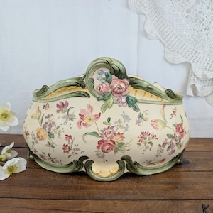 Peut inclure: Une jardinière murale en céramique de couleur crème ornée d'un motif floral coloré composé de fleurs roses, jaunes et violettes. Elle présente un bord festonné décoratif avec une bordure verte et un relief floral en haut au centre.