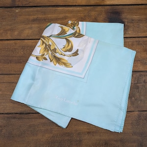 Peut inclure: Un foulard en soie bleu clair avec un motif floral décoratif doré et blanc. Le foulard porte le texte "Guy Laroche" dans le coin. Le foulard est plié sur une surface en bois.