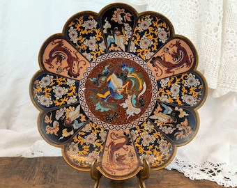 Peut inclure: Une grande assiette décorative ronde et festonnée avec un motif en émail cloisonné noir, bleu, or et rouge. L'assiette présente un motif central de phénix et de dragons, entouré de motifs floraux. L'assiette est sur un support en métal doré.